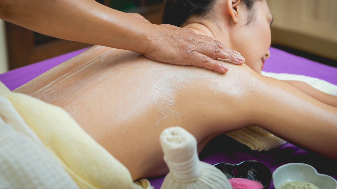 Una persona recibe un masaje de espalda con aceite en un spa, con compresas de hierbas y cuencos cerca.