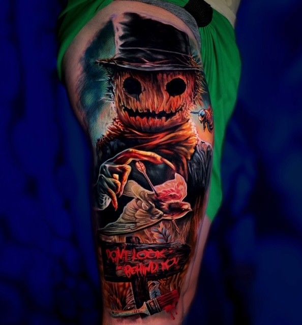 Buntes Tattoo einer vermummten Vogelscheuche mit leuchtenden Augen und aufgenähtem Grinsen auf einem Oberarm