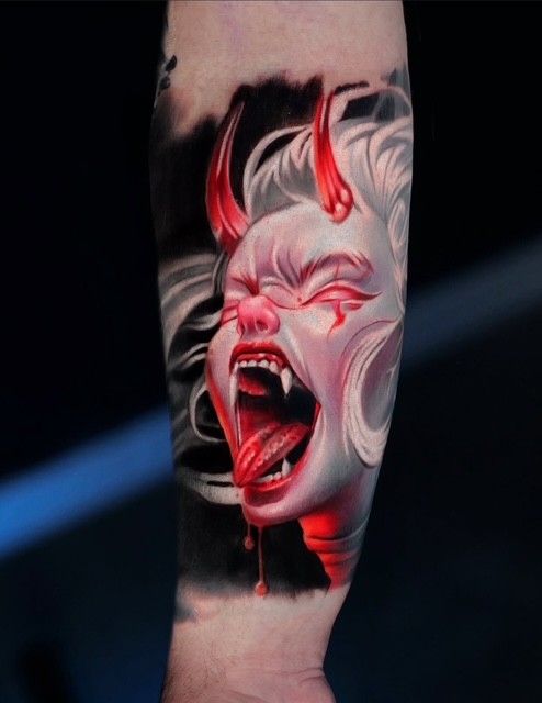 Rot-weißes dämonisches Gesichtstattoo mit Hörnern, offenem Mund und intensivem Ausdruck auf einem Arm