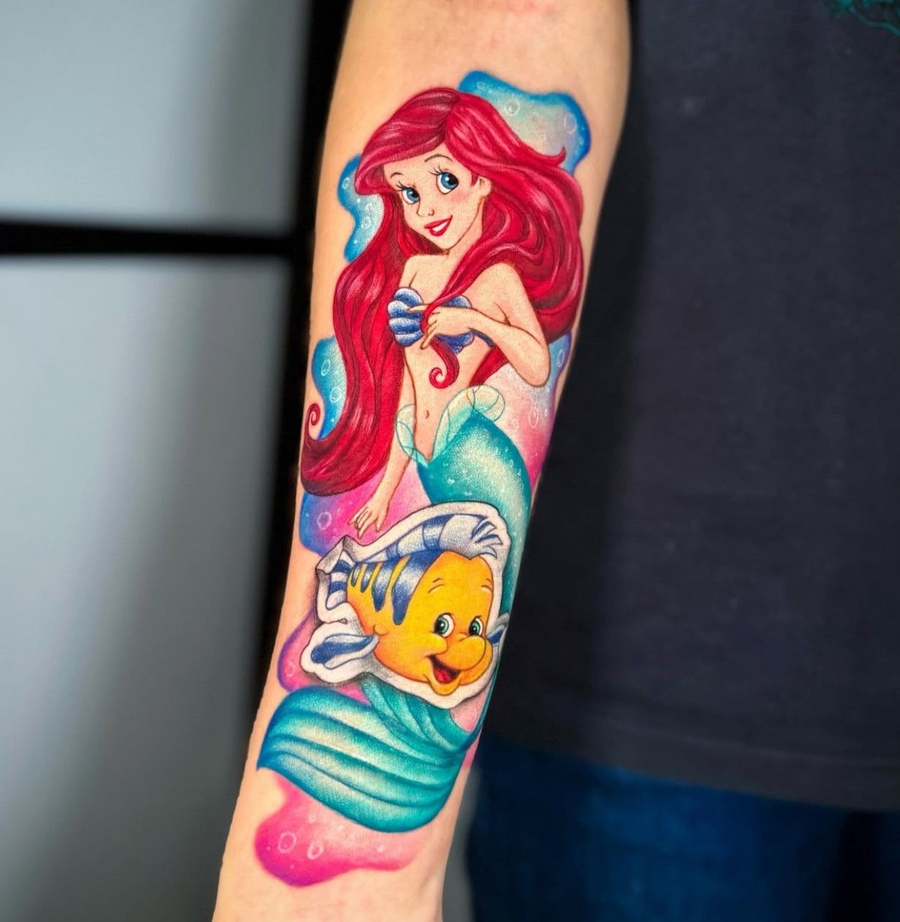 Eine Person hat eine Tätowierung von Arielle und Fabius auf dem Unterarm.