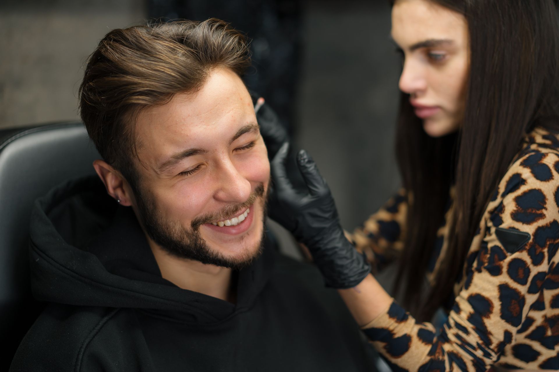 Piercerin kontrolliert im Rahmen der Piercing Pflege das frisch gestochene Ohrpiercing eines lächelnden jungen Mannes. Piercerin kontrolliert im Rahmen der Piercing Pflege das frisch gestochene Ohrpiercing eines lächelnden jungen Mannes.
