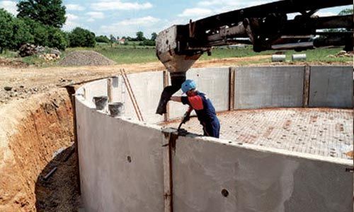 Un ouvrier du bâtiment installe un segment de mur en béton préfabriqué pour une structure circulaire. Des fouilles sont effectuées autour de celle-ci.