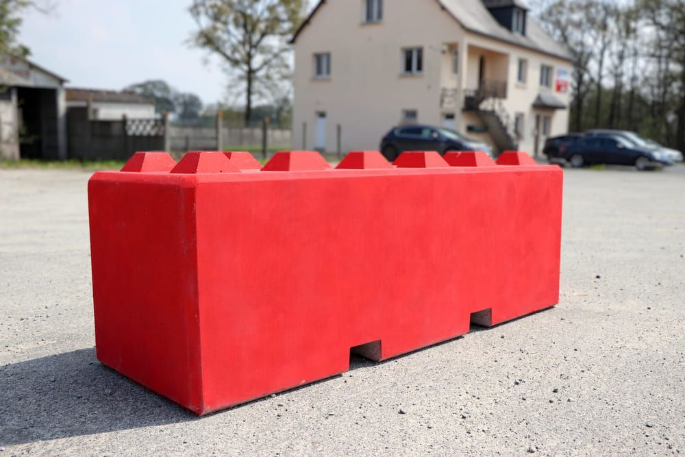 Bloc de béton rouge ressemblant à une brique Lego, posé sur une surface de gravier, avec un bâtiment et des voitures en arrière-plan.