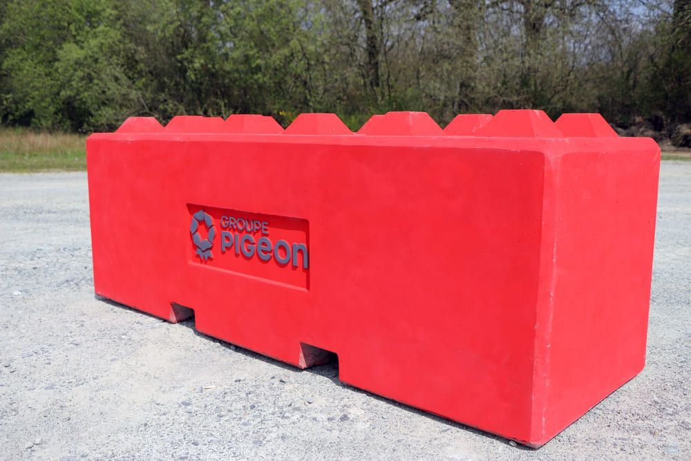 Bloc de plastique rouge rectangulaire avec tenons et encoches en creux, semblable à des Lego ; logo « Lego Pigeon ». À l’extérieur, sur le trottoir.