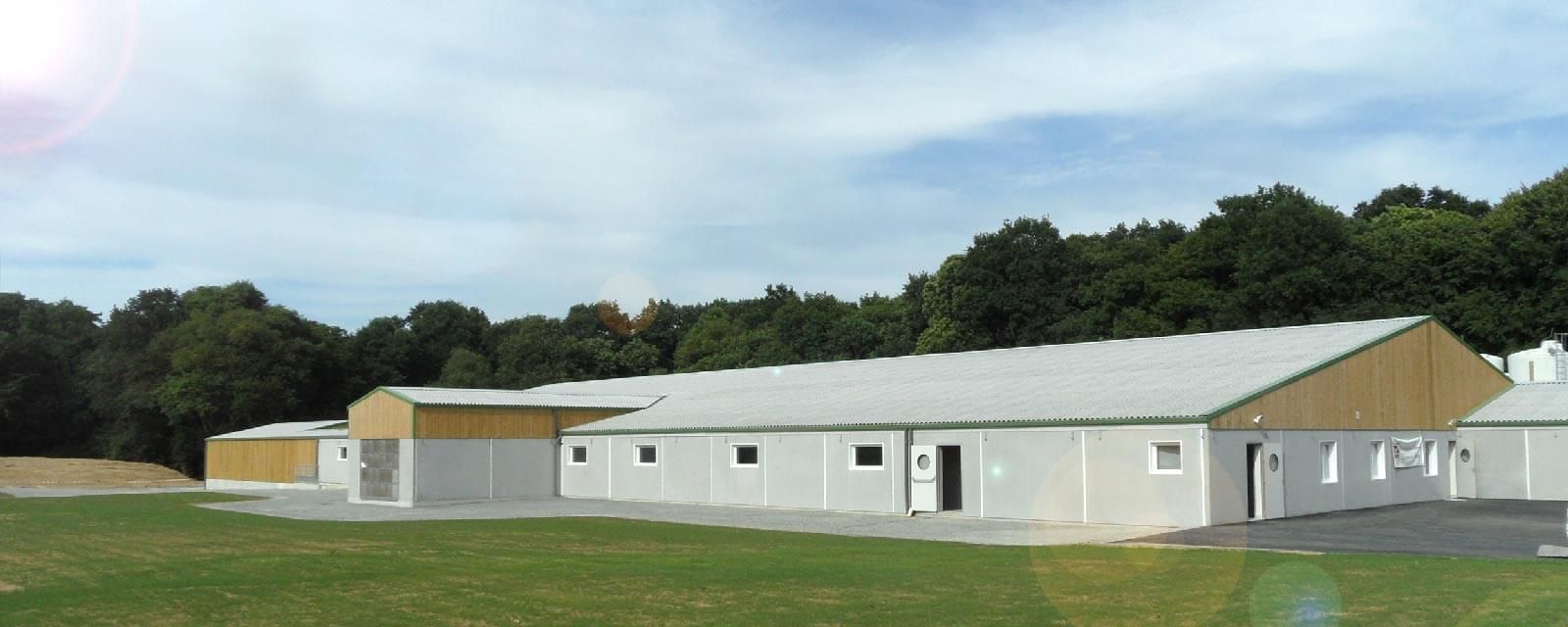 Un long bâtiment clair au toit en pente. La structure se détache sur un champ verdoyant, avec des arbres en arrière-plan.