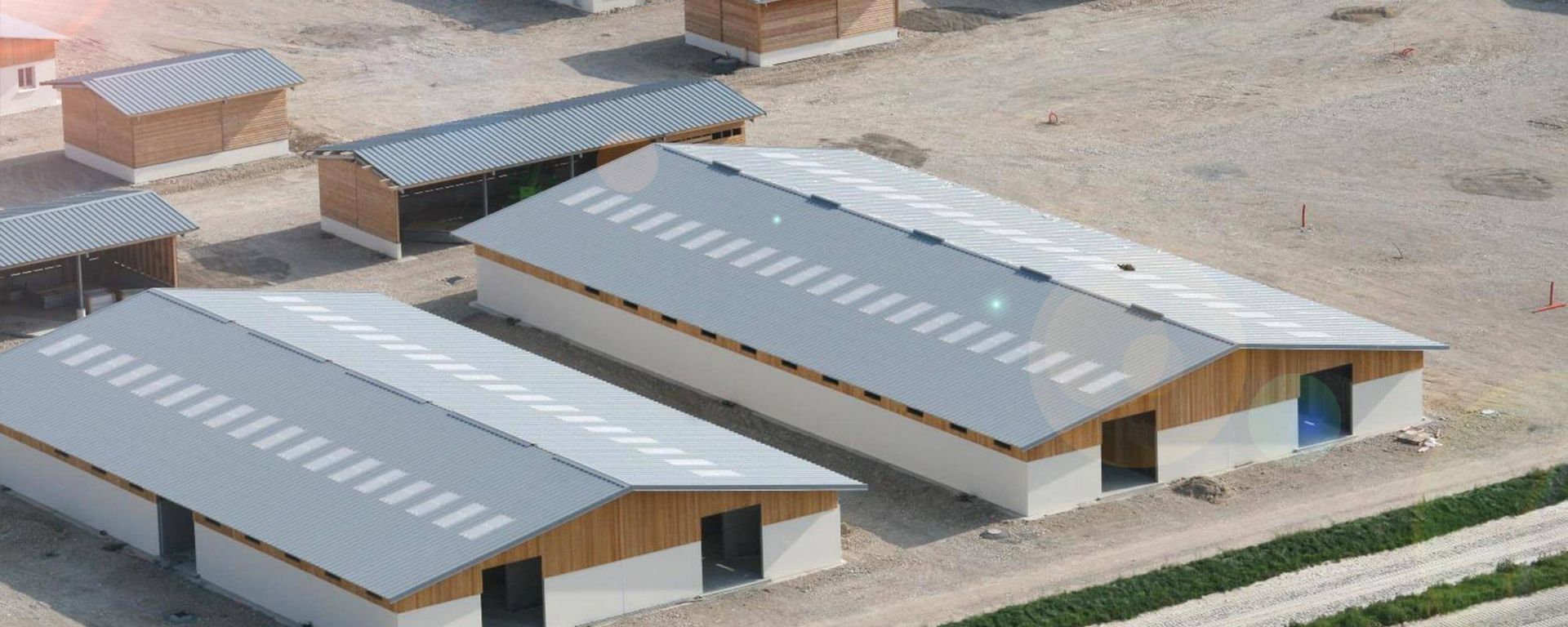 Vue aérienne de plusieurs bâtiments rectangulaires aux toits métalliques et aux éléments en bois, sur un terrain sablonneux.