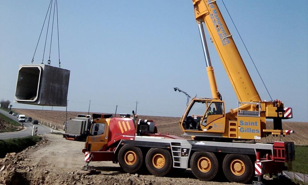 Une grue jaune soulève une grande structure en béton à côté d'un chemin de terre.