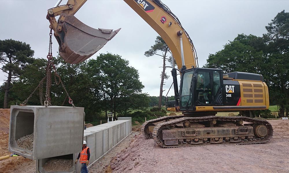 Une pelleteuse pose un grand ponceau en béton sur un chantier. Un ouvrier se tient à proximité.