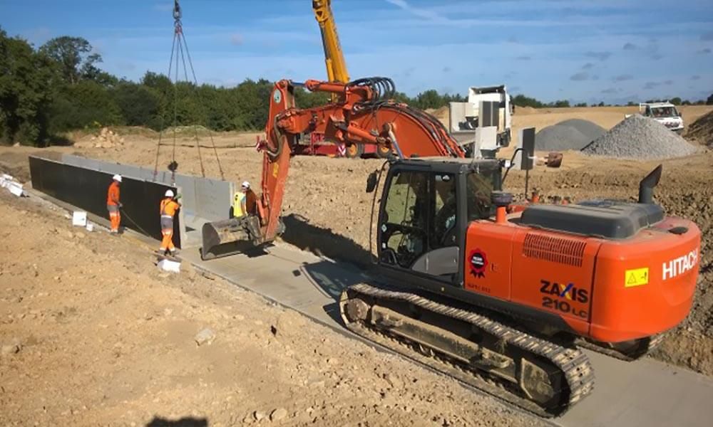 Chantier : Une excavatrice et une grue soulèvent une barrière en béton ; des ouvriers portent des gilets orange ; de la terre, du gravier et des arbres sont visibles en arrière-plan.