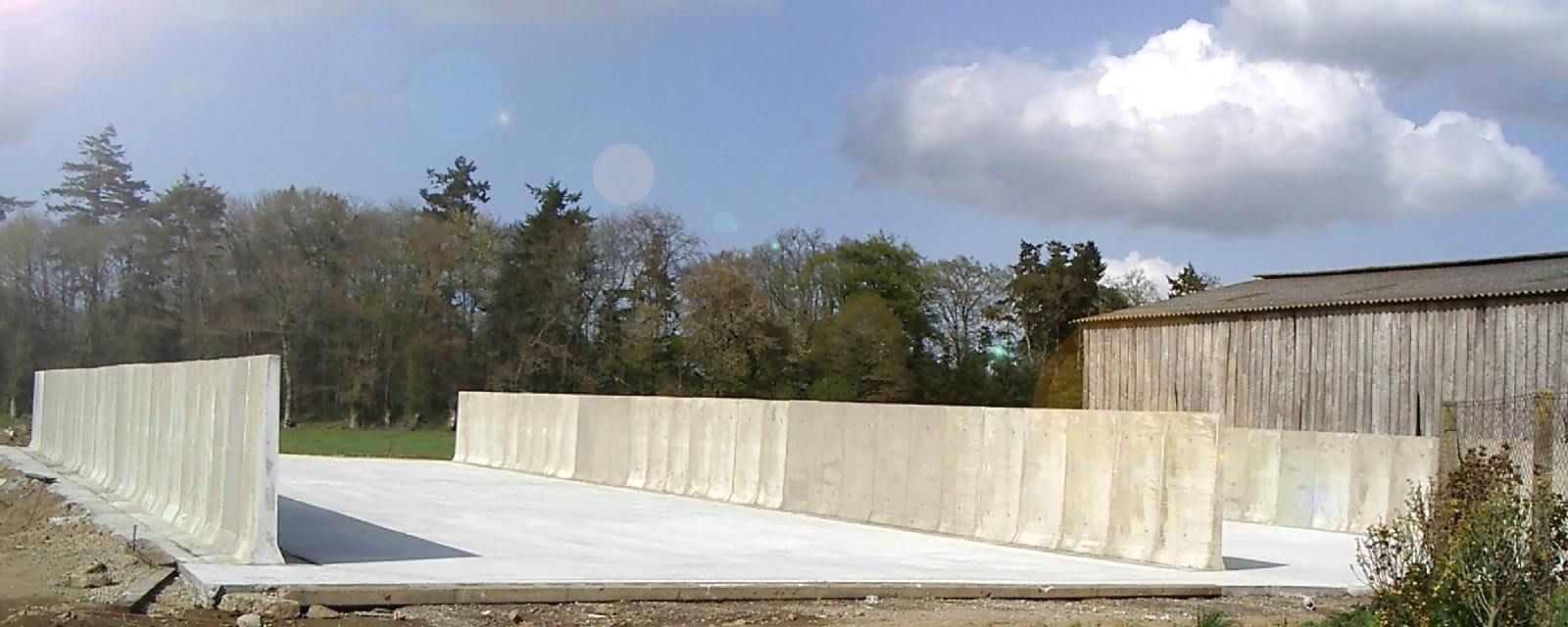 Structure en béton avec murs et sol en béton, se détachant sur un fond d'arbres et d'un bâtiment en bois, sous un ciel bleu.