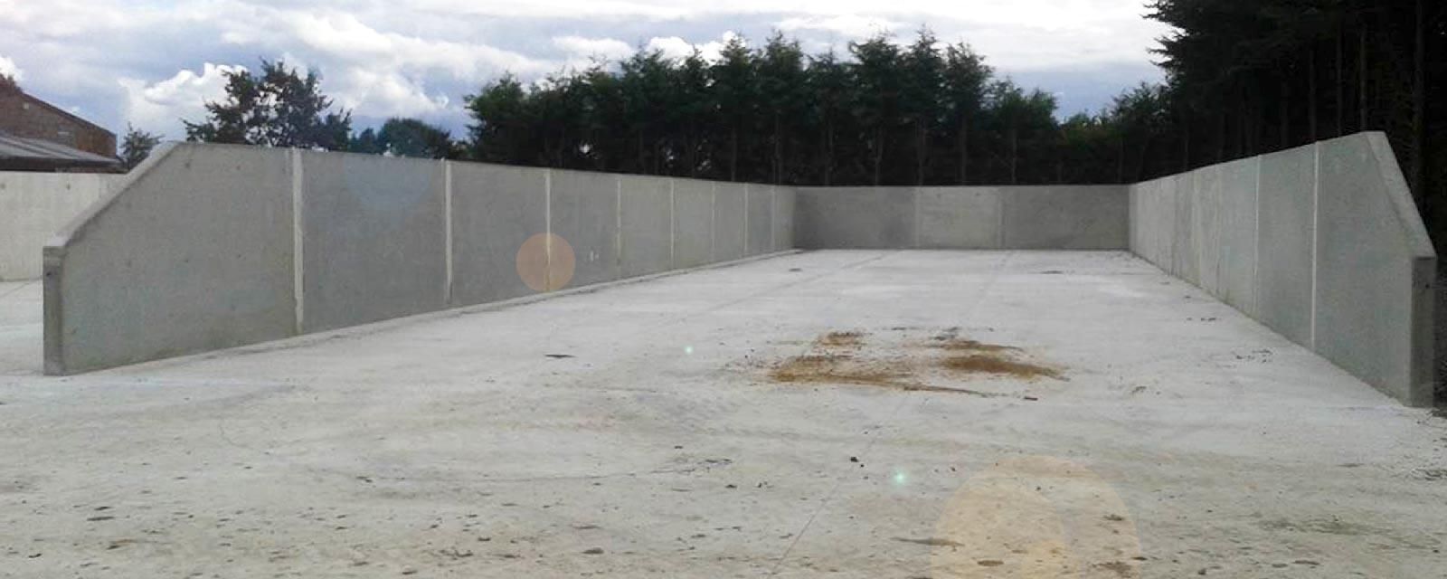 Des murs de béton entourent une zone bétonnée vide, avec des arbres en arrière-plan sous un ciel nuageux.