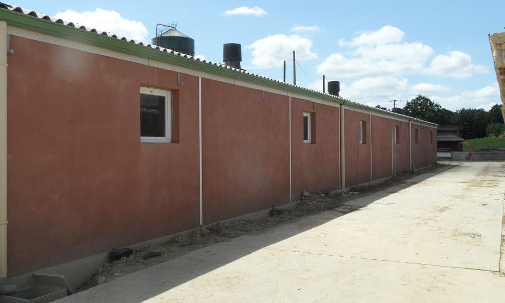 Long bâtiment brun rougeâtre aux petites fenêtres, bordant un chemin pavé, sous un ciel bleu.