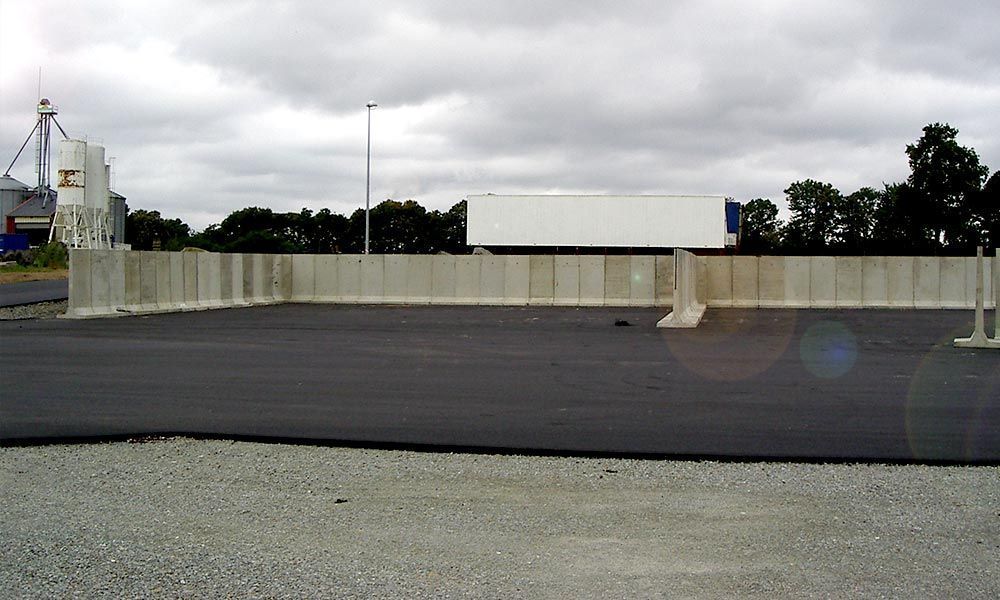Terrain en asphalte noir bordé d'un mur en béton, avec un semi-remorque en arrière-plan sous un ciel nuageux.