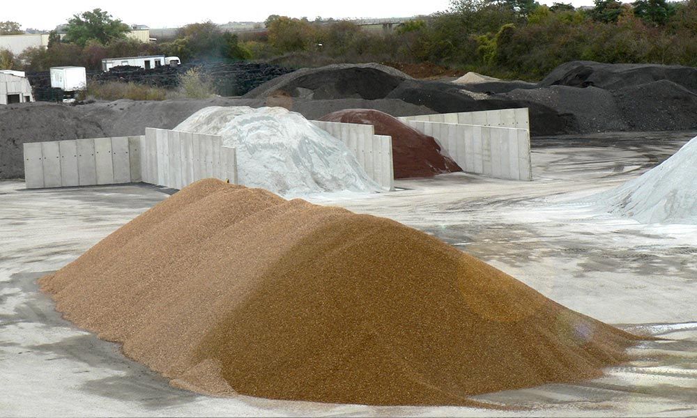 Des tas de matériaux de construction : gravier beige, agrégats blancs, rouges et gris foncé, et barrières en béton. À l’extérieur.