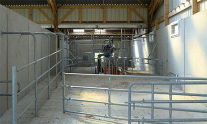 Intérieur d'une grange avec portails et stalles métalliques. Une machine se trouve au centre. La lumière du soleil pénètre dans la structure.