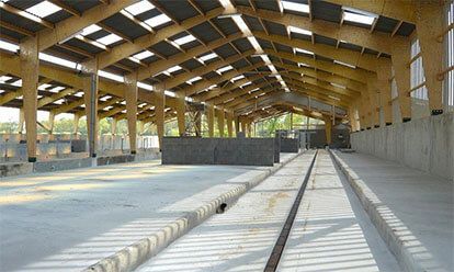 Grand bâtiment ouvert avec une charpente en bois. Sol et murs latéraux en béton, puits de lumière.
