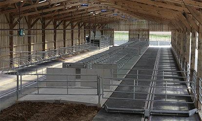 Intérieur d'une grange avec stalles, rambardes métalliques et large allée centrale. Des poutres en bois soutiennent le toit.