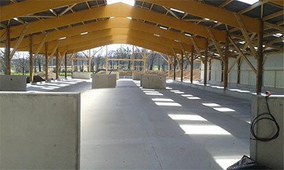 Bâtiment agricole à ossature bois et sol en béton, vide. La lumière filtre à travers le toit ouvert.