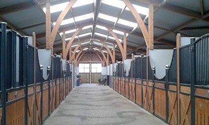 Intérieur d'une écurie avec des boxes bordant une allée sablonneuse, poutres en bois au-dessus.