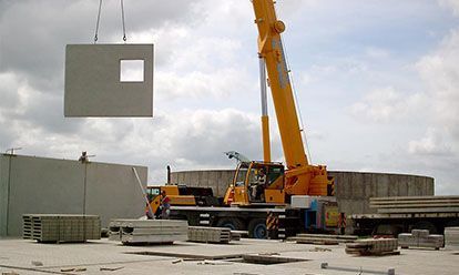 Grue soulevant un panneau mural en béton avec une fenêtre, sur un chantier de construction.