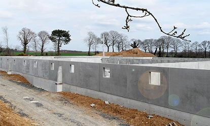 Murs de fondation en béton en construction dans un cadre rural, avec des arbres et un ciel nuageux en arrière-plan.