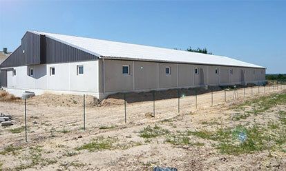 Long bâtiment rectangulaire aux murs clairs, au toit blanc et à la façade en bois, sous un ciel bleu limpide.