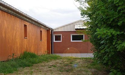Deux bâtiments au revêtement orange, reliés par un chemin ; de l'herbe verte et des buissons les entourent.