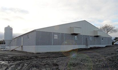 Grand bâtiment industriel gris avec silo et terrain environnant en terre.