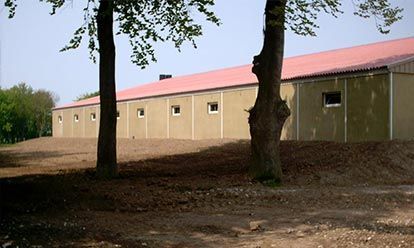 Bâtiment long et beige avec un toit rouge, de petites fenêtres et des arbres à l'avant, sur un terrain légèrement surélevé.