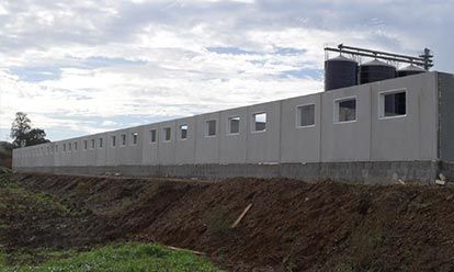Long bâtiment en béton gris avec de petites fenêtres carrées, deux silos au sommet et de la terre devant.
