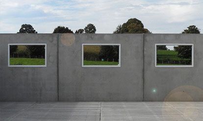 Mur gris percé de trois ouvertures rectangulaires reflétant un champ verdoyant et des arbres sous un ciel nuageux.
