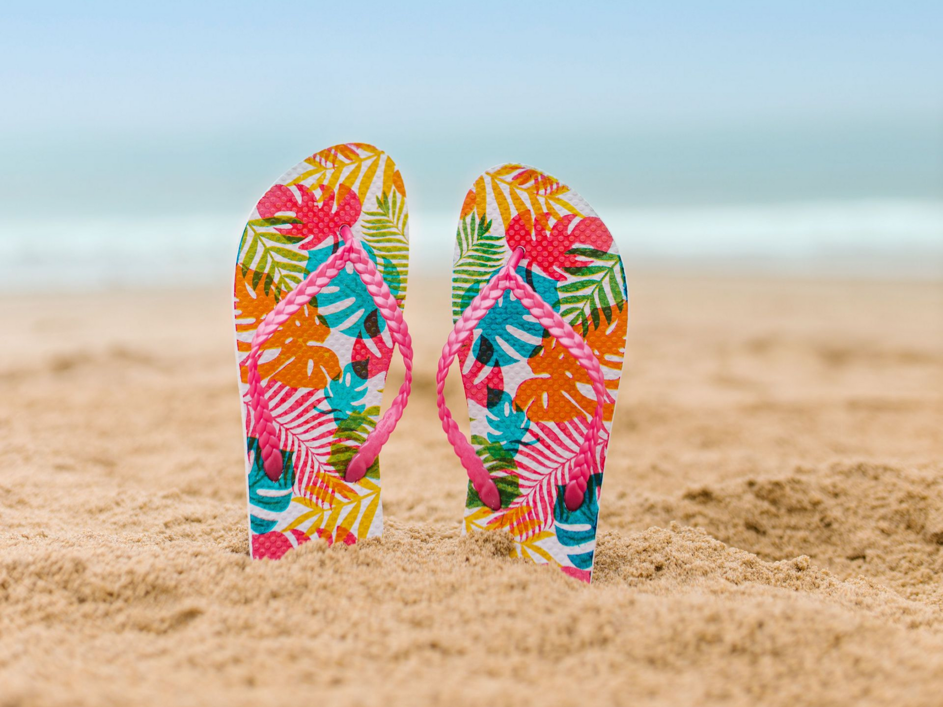 Einschränkungen in der Honorierung unserer Leistungen Bunte, blumengemusterte Flip-Flops stehen aufrecht an einem Sandstrand, im Hintergrund ist das Meer verschwommen zu sehen.