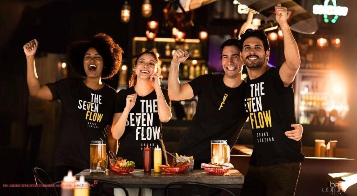 Cuatro personas con camisas negras en un bar animan con los brazos en alto. Se ve el logo de 