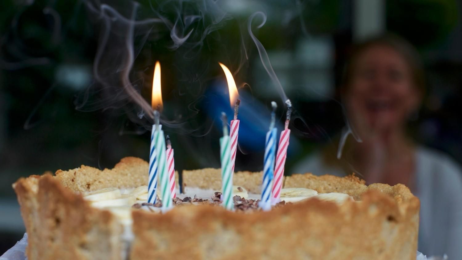 Pastel con velas de cumpleaños encendidas; una persona borrosa sonríe en el fondo.