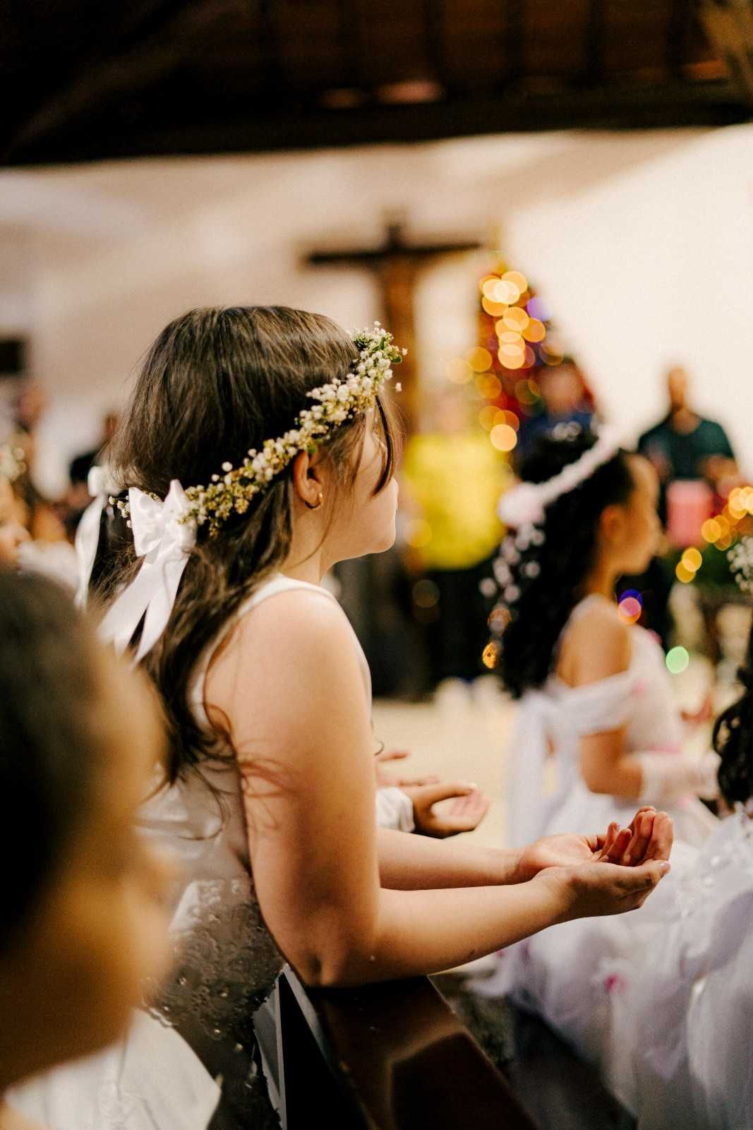 Niña con vestido blanco y corona de flores, manos entrelazadas, en una ceremonia.