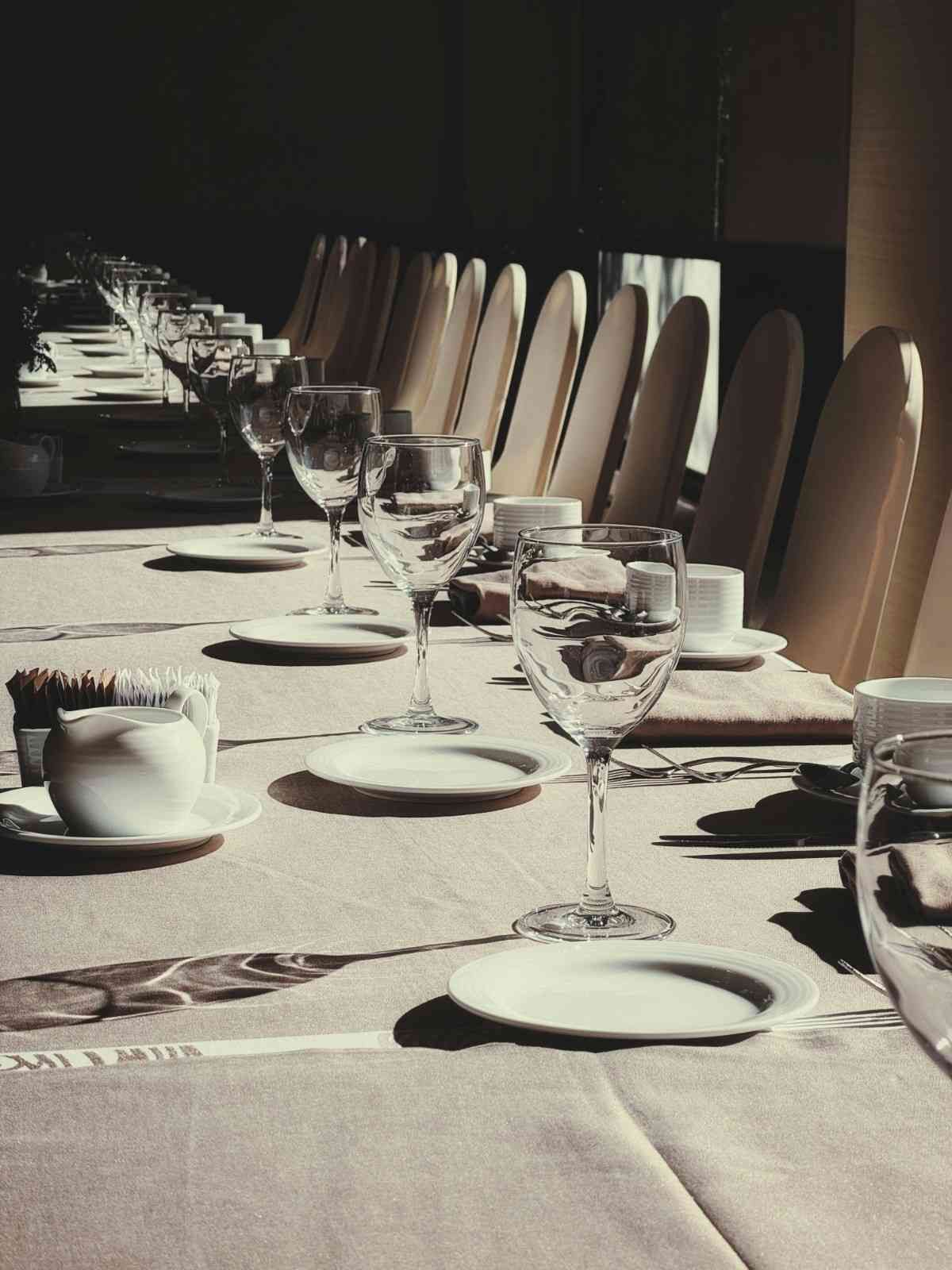 Una mesa puesta con vasos, platos y sillas. La luz del sol proyecta sombras sobre un mantel blanco.