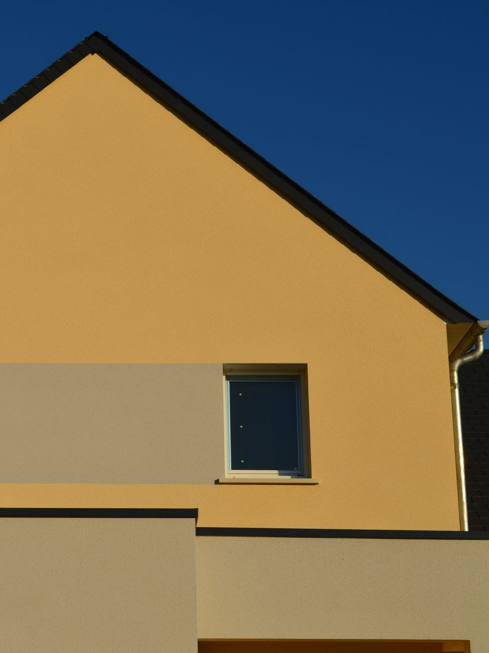 Maison jaune avec une petite fenêtre carrée et une bande grise contrastante.