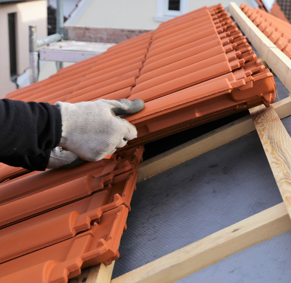 Une personne portant des gants installe des tuiles orange sur une charpente en bois.