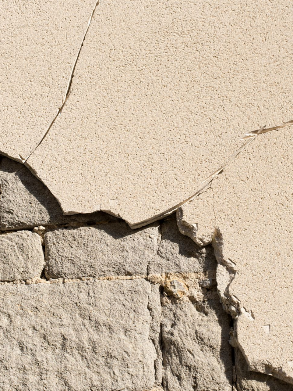 Enduit fissuré sur un mur en pierre. L'enduit est beige clair et la pierre est grise.