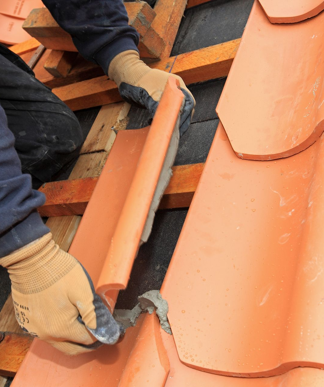 Une personne portant des gants installe des tuiles de toit en terre cuite sur une charpente en bois.