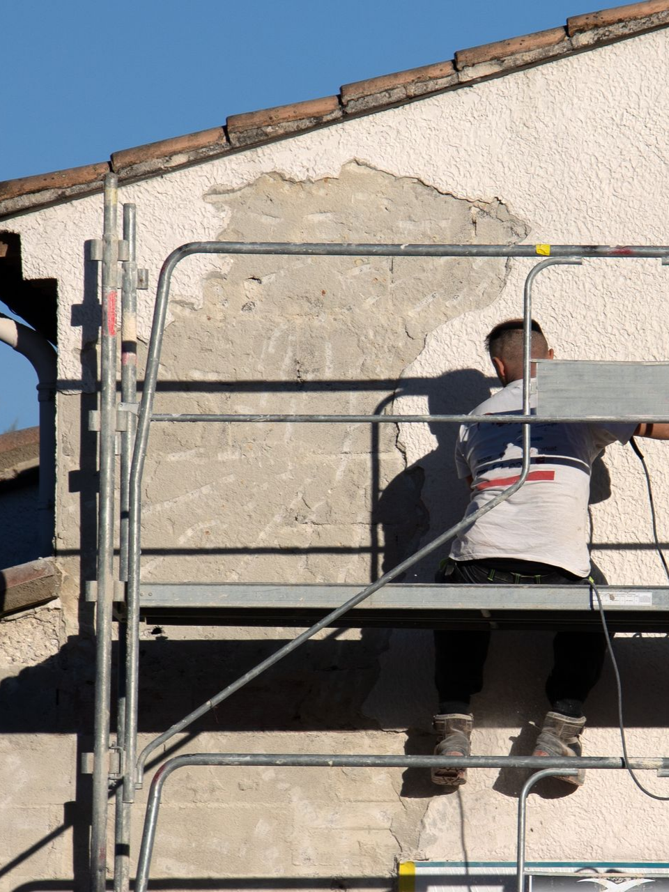 Une personne sur un échafaudage répare un mur en stuc ; une partie de la surface endommagée est visible.