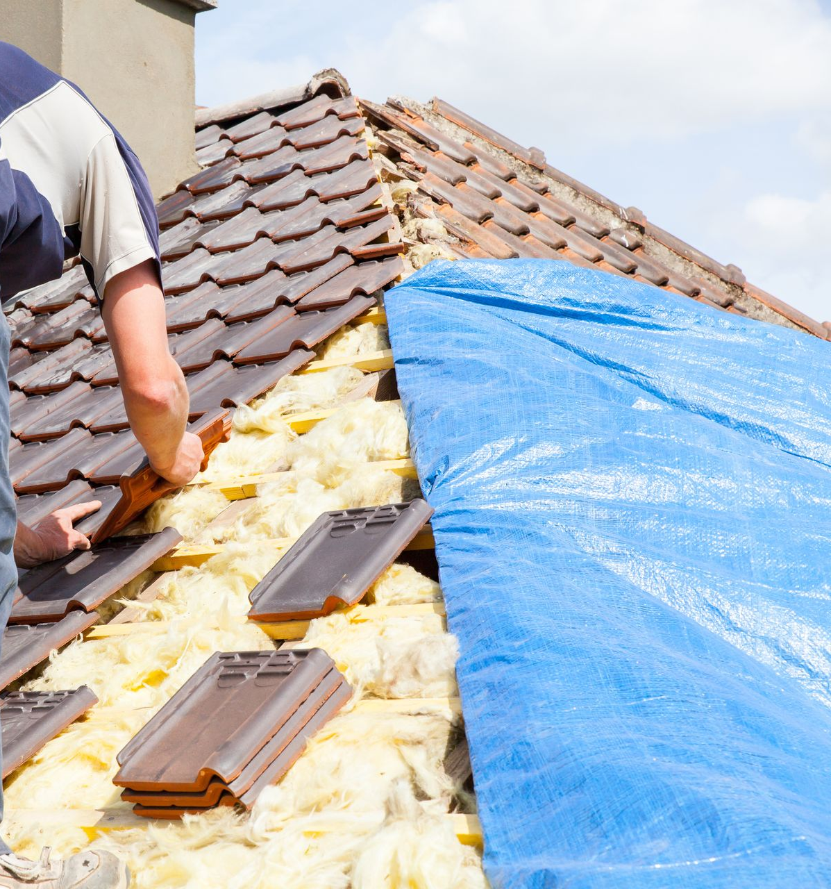 Un homme pose des tuiles sur le toit d'une maison. Une bâche bleue recouvre la partie exposée.