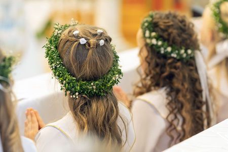 Zwei Kinder mit Blumenkränzen sitzen an einem Tisch, von hinten gesehen, in einer hellen, festlichen Umgebung