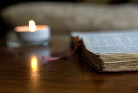 Ein von Kerzenlicht erhelltes, aufgeschlagenes Buch liegt auf einem Holztisch, warme Lichtreflexe spiegeln sich im Hintergrund.