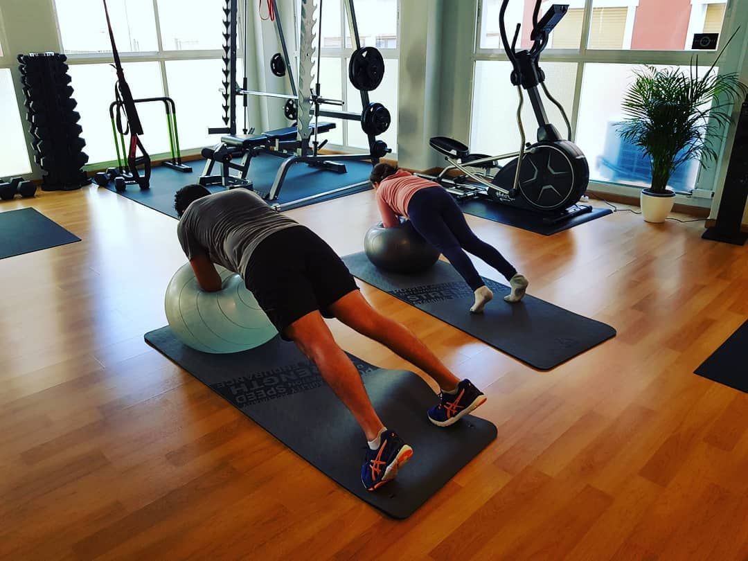Dos personas haciendo ejercicio en un gimnasio: planchas sobre pelotas de ejercicio, colchonetas en el suelo, equipo de pesas en el fondo.