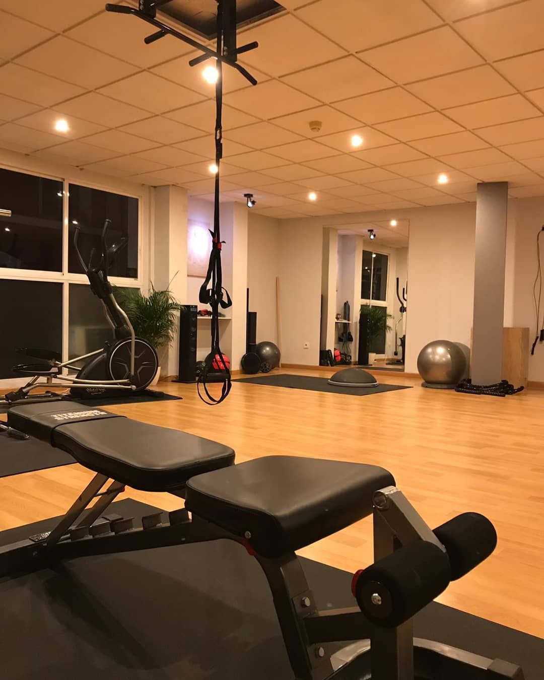 Interior de gimnasio con máquinas de ejercicio sobre suelo de madera. Incluye banco de pesas, elíptica, correas TRX y pelotas de equilibrio.