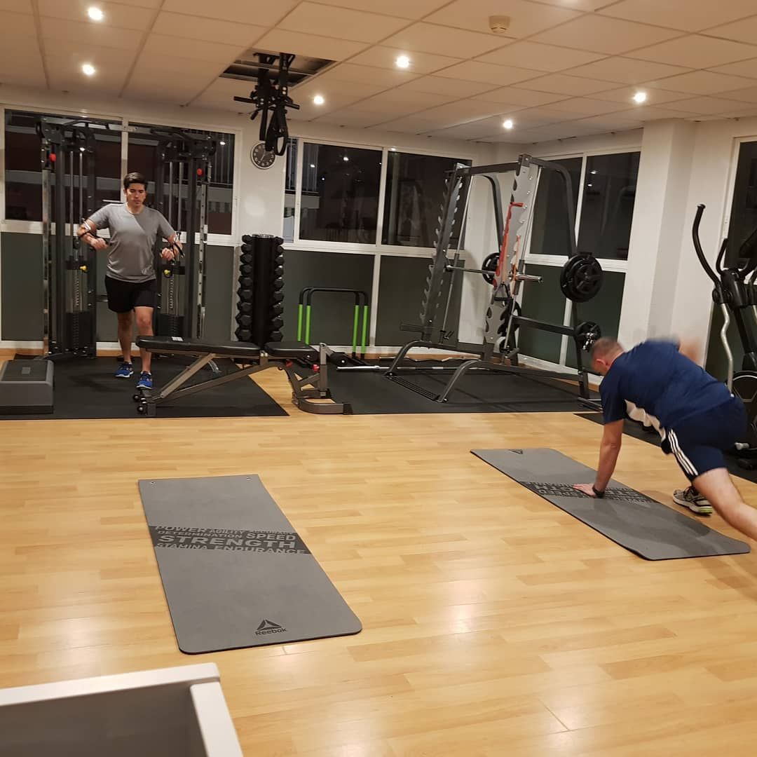 Dos personas haciendo ejercicio en un gimnasio: una corre, la otra hace plancha sobre una colchoneta. Pesas y aparatos al fondo.