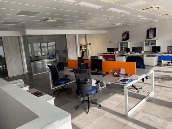 Oficina moderna de planta abierta con escritorios, sillas negras, separadores naranjas y ordenadores bajo luces de techo brillantes.