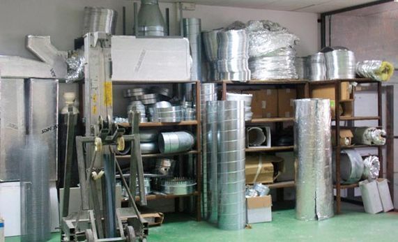 Trastero con conductos metálicos apilados, ventiladores y estanterías con piezas industriales sobre un suelo verde.