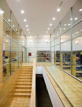 Interior de biblioteca moderna con paredes de cristal, suelos de madera y una escalera central que da acceso a un nivel inferior.